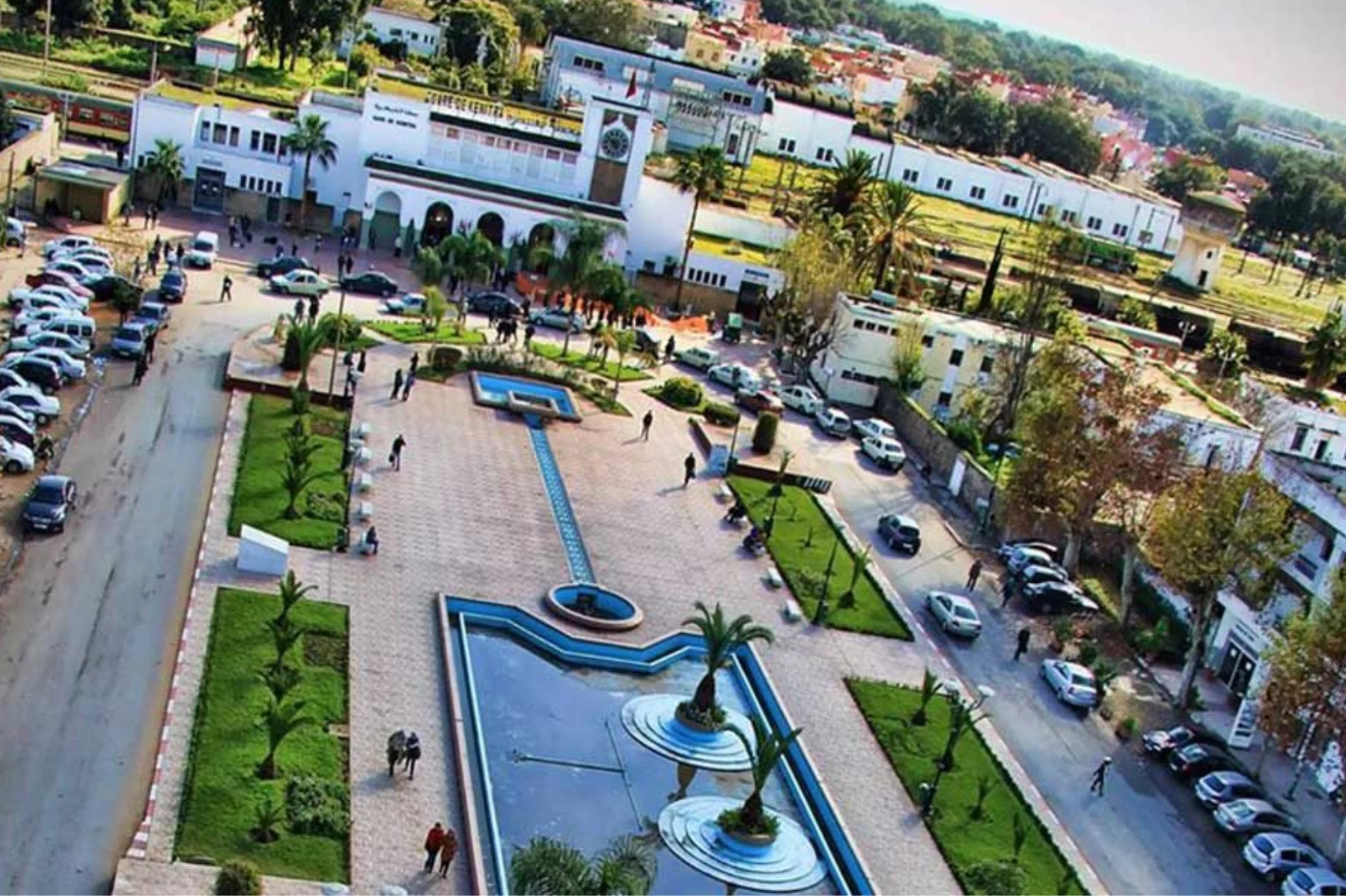 Place aménagée avec fontaine devant une gare blanche.