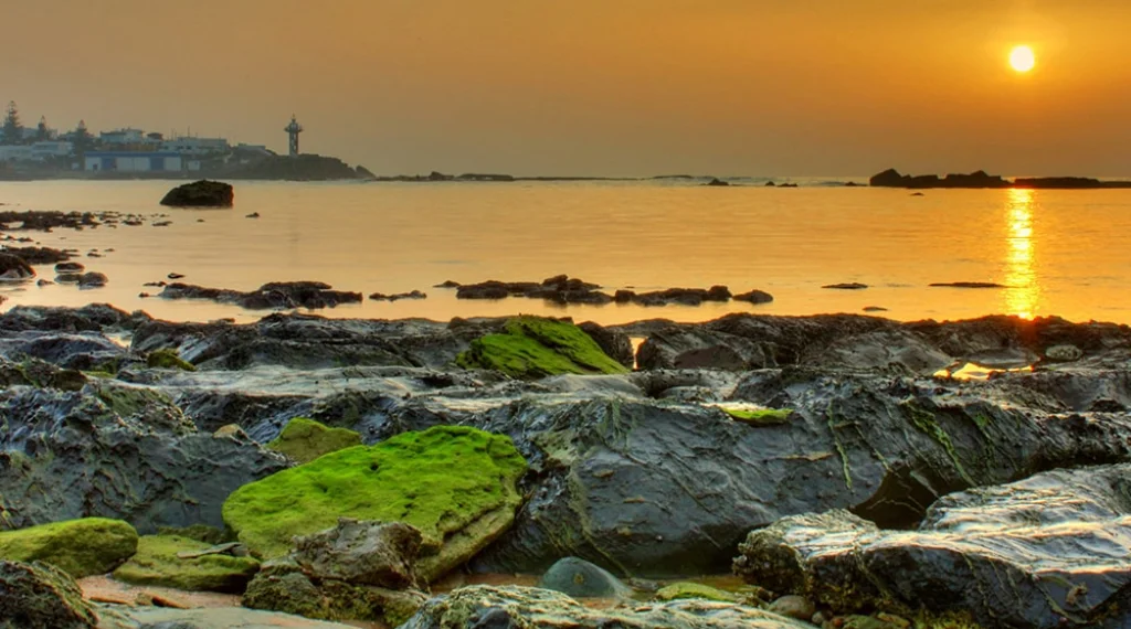 Coucher de soleil doré sur une plage rocheuse avec un phare au loin.