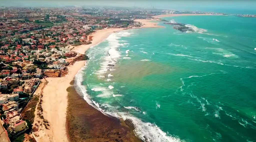 Vue aérienne de la côte et des plages de Mohammedia au Maroc, montrant une mer turquoise avec des vagues et un quartier résidentiel en bord de mer.